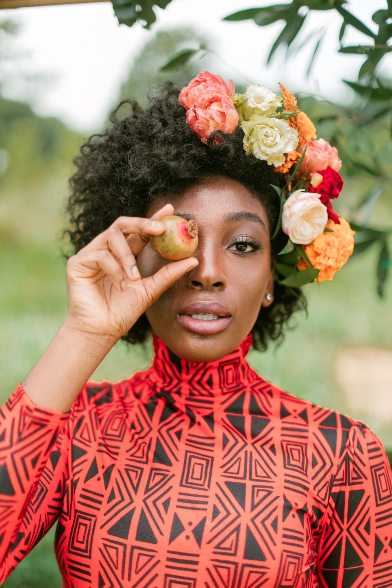 A woman wearing a colorful floral crown holds a small apple over one eye