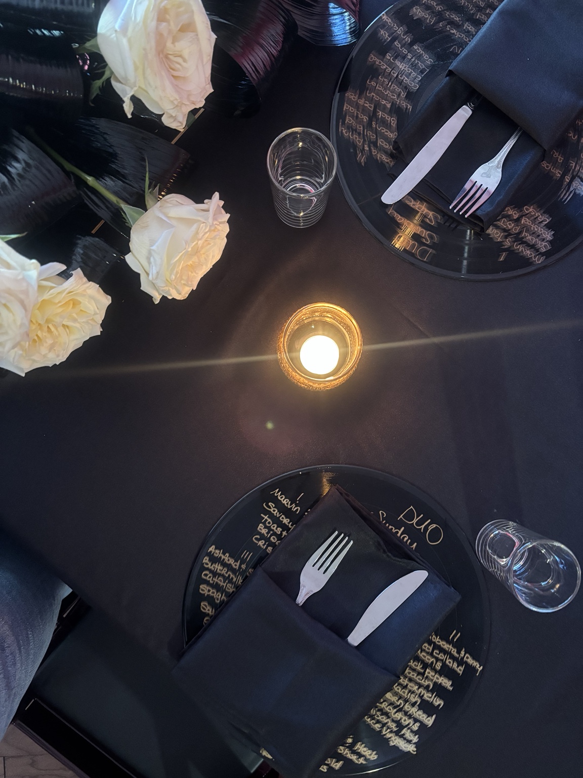 A candle glowing among vinyl menus, black napkins, and white roses at Sunday Supper