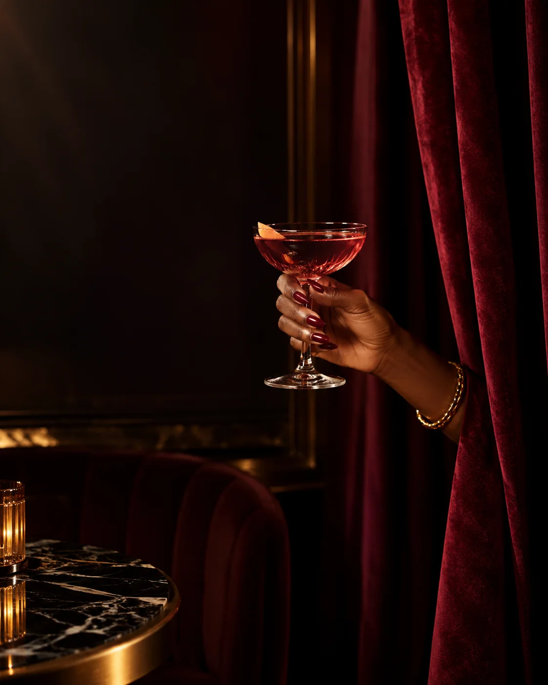 A manicured hand presents a garnet cocktail through burgundy velvet curtains in a candlelit salon